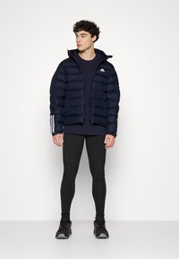 Polstret navyjakke med hætte og lynlåsfront, med hvide Adidas-logoer og tre striber på ærmerne, parret med sorte leggings og sko.