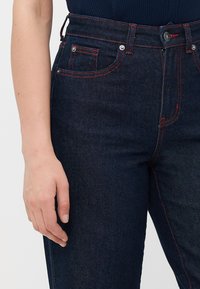 Venstre hånd hviler ved siden af mørkeblå jeans med rød syning og sølvnitter, iført en mørk top.