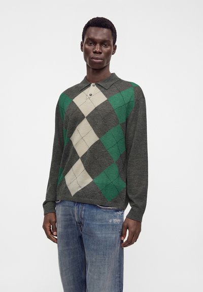 Млад мъж, носещ тъмнозелен и сив аргайл пуловер с яка и сини избелели дънки, стои пред обикновен светъл фон.