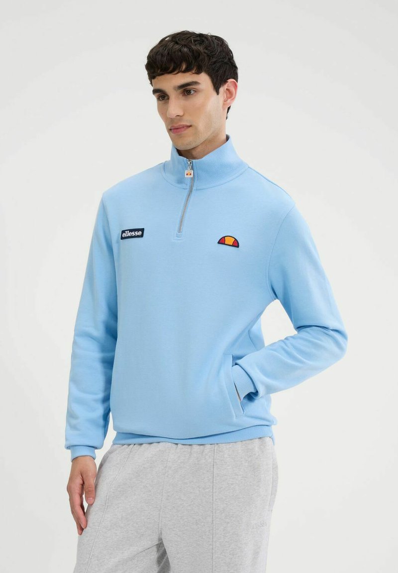 Sudadera con cremallera de color azul claro, hecha de un tejido suave. Presenta un cuello alto, puños elásticos y un parche con el logo en el pecho.