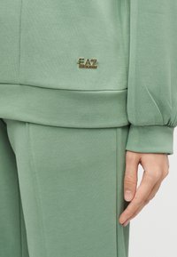 Κοντινό πλάνο σε σετ ανοιχτοπράσινης φανέλας και παντελονιού με χρυσό λογότυπο EA7 Emporio Armani στο τελείωμα της φανέλας, που δείχνει το μανίκι σε άνετη εφαρμογή και ένα χέρι.