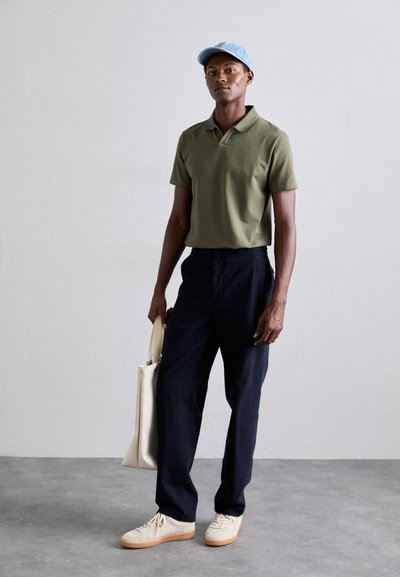 Olivgrön polo-shirt, marinblå byxor, beige sneakers och ljusblå keps. Modellen håller en krämfärgad tote-väska. Mjuka texturer, avslappnad design.