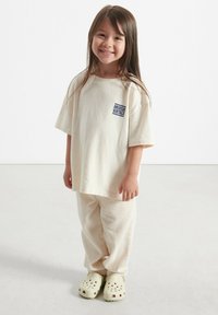 T-shirt beige surdimensionnée avec logo "American Vintage", pantalon beige côtelé et sabots vert clair avec des trous de ventilation.