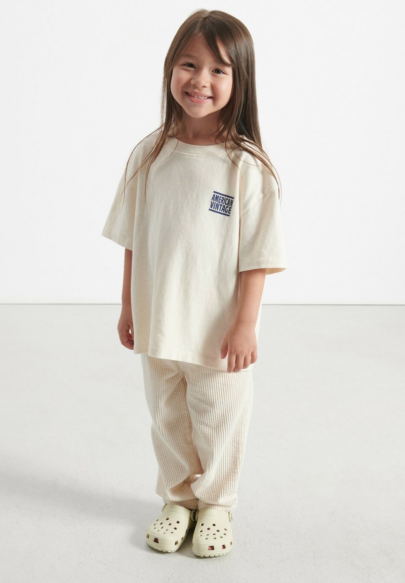 T-shirt beige surdimensionnée avec logo "American Vintage", pantalon beige côtelé et sabots vert clair avec des trous de ventilation.