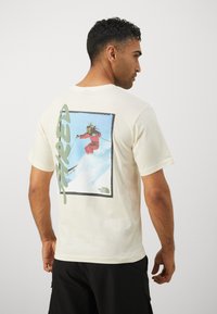 Cream short-sleeve t-shirt featuring a skier graphic on the back with green text "FIRED," and "The North Face" logo in the corner.