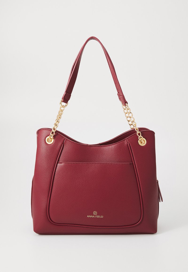 Sac à main en simili cuir rouge avec une finition texturée, accent en chaîne dorée et un grand poche frontale. Logo doré à l'avant.