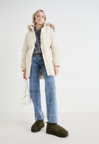Pufferjack in crème met een bont gevoerde capuchon, grijs sweater, blauw gestreepte jeans met studs en olijfgroene platformlaarzen. Witte handtas.