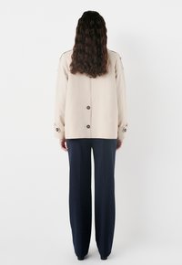 Giacca outerwear beige con vestibilità rilassata, dotata di chiusura a bottoni, dettagli sui polsini e pantaloni a gamba larga blu navy, vista da dietro.
