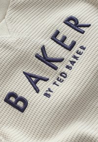Tissu texturé couleur crème avec un motif en grille comportant un texte brodé bleu marine : "BAKER par TED BAKER," affiché de manière proéminente.