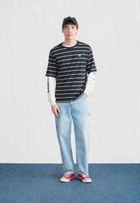 Νεαρός άνδρας με μαύρο καπέλο, μαύρο και λευκό ριγέ πουκάμισο πάνω από λευκά μανίκια, ανοιχτό μπλε τζιν και κόκκινα αθλητικά παπούτσια στέκεται σε μπλε χαλί.