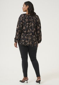 Sort blomstret bluse med gathered ryg, der har lange ærmer og en løs pasform, parret med mørke skinny jeans og sorte hælede sandaler.