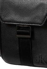 Fekete, texturált bőrtáska fedél fekete pánttal és fém csattal, a jobb alsó sarokban finoman dombornyomott "ck" logóval.