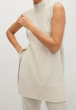 Tunique sans manches en maille beige à col montant portée avec un pantalon en maille assorti, personne avec une main sur la hanche et l'autre main levée près de la poitrine.