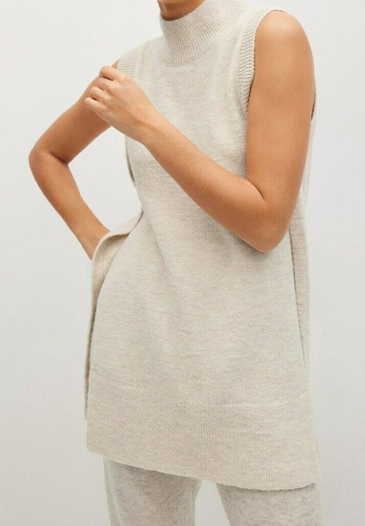 Tunique sans manches en maille beige à col montant portée avec un pantalon en maille assorti, personne avec une main sur la hanche et l'autre main levée près de la poitrine.