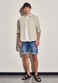 Světle zelená košile s dlouhým rukávem přes bílý tričko, doplněná o ohrnuté džínové šortky a černé sandály na texturované podlaze.