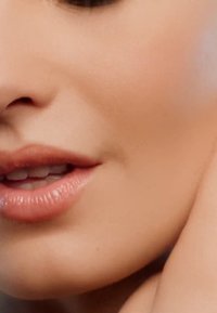 Gros plan sur des lèvres lisses et légèrement brillantes, d'une teinte naturelle, mettant en avant un sourire subtil et une texture de peau douce, accentués par un éclairage doux.