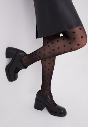 Schwarze klobige Schuhe mit Absatz, runder Spitze und Riemen, kombiniert mit schwarzen, transparenten Strumpfhosen mit schwarzen Punkten vor weißem Hintergrund.