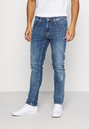 Vaqueros de denim azul con un corte ajustado, que presentan un detalle desgastado en una pierna, cinco bolsillos y un cierre clásico de botón y cremallera.