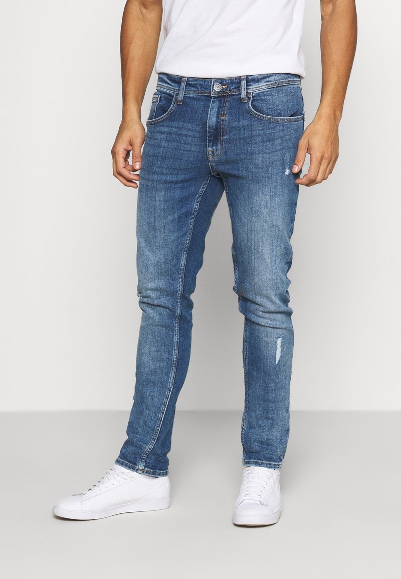 Vaqueros de denim azul con un corte ajustado, que presentan un detalle desgastado en una pierna, cinco bolsillos y un cierre clásico de botón y cremallera.