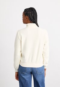 Cream-colored fleece sweatshirt with a high collar, ribbed cuffs, and a fitted hem, paired with light blue denim jeans.
