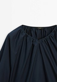 Blouse bleu marine avec encolure ronde froncée et petite fermeture éclair au centre devant, montrant le haut de la poitrine et les épaules.