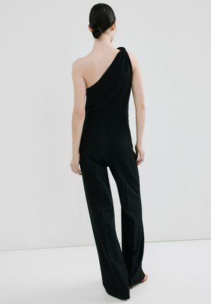 Femme aux cheveux foncés coiffés en chignon bas, portant une combinaison noire à une épaule avec des jambes larges, debout devant un fond gris clair.