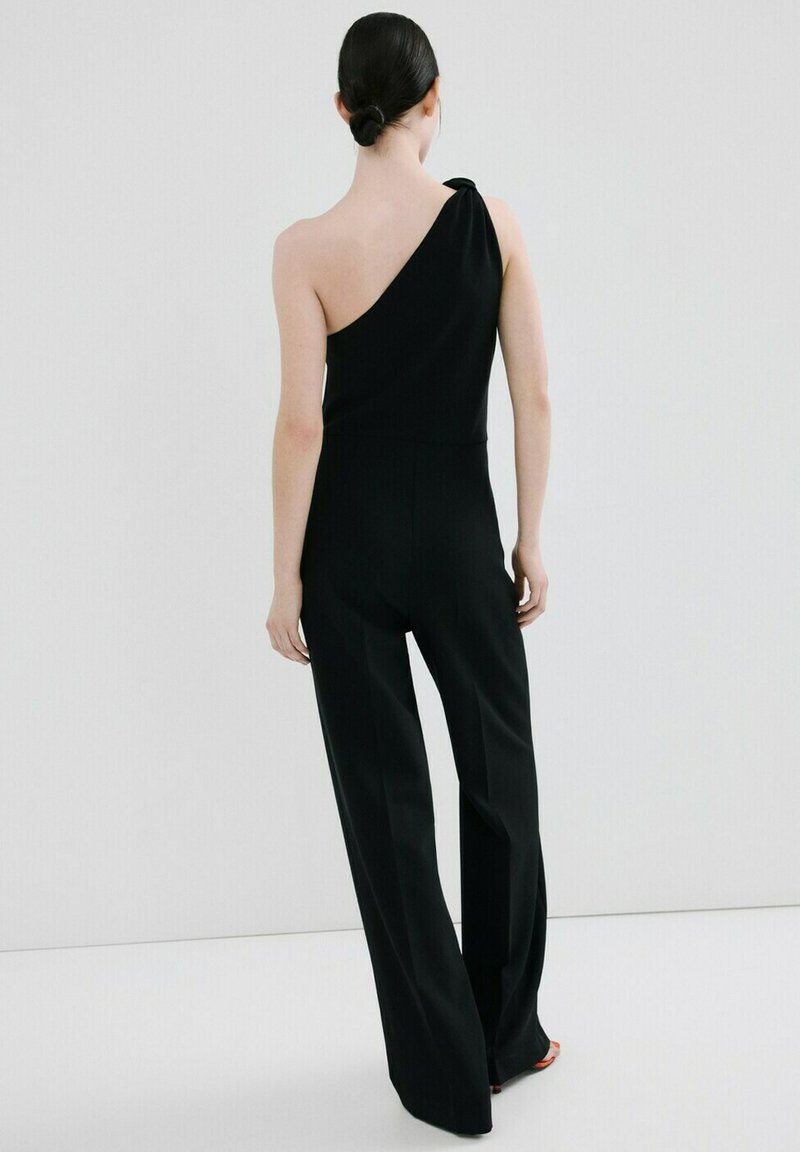 Žena s tmavými vlasy staženými do nízkého drdolu, oblečená v černém overal s jedním ramenem a širokými nohavicemi, stojící na světle šedém pozadí.