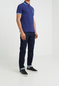 Polo shirt marine à manches courtes, avec des accents rayés bleu clair, design brodé sur la poitrine, associé à un jean en denim foncé et des baskets noires.