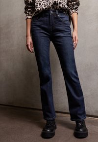 Jeans de mezclilla azul oscuro con corte recto, que presentan un diseño de cinco bolsillos y costuras sutiles. Combinados con botas negras gruesas de cordones.