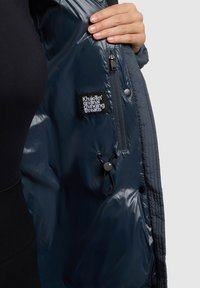 Tamsiai mėlyna pūkinė striukė su blizgia danga, pasižyminti užtrauktuku su kišeniu ir raišteliu. Etiketėje užrašyta "Khujo Original Changing Threads."