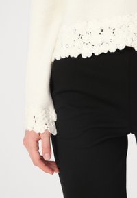 Pull en tricot blanc avec un ourlet en dentelle florale texturée, porté sur un pantalon noir ajusté, mettant en valeur des embellissements floraux complexes.