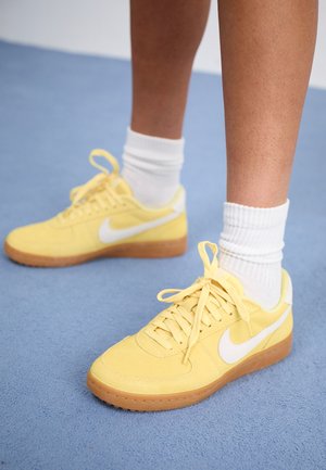 Jambes avec des chaussettes blanches et des baskets Nike jaunes avec des logos swoosh blancs sur un sol moquetté bleu.