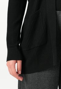 Handen vilar vid den svarta ribbade cardiganens ficka och de grå herringbone-mönstrade byxorna, vilket visar tygtextur och kläddetaljer.
