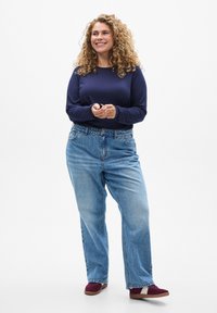Navy long-sleeve shirt, relaxed-fit light blue jeans, and burgundy sneakers. Curly hair, standing with hands clasped. Plain white background.