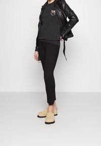 Giubbotto di pelle nera sopra un felpa scura con logo, abbinato a pantaloni neri slim-fit e zoccoli in suede crema con suole nere.