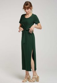 Robe vert foncé, longueur genoux, en tissu doux, avec un lien à la taille et des fentes sur les côtés ; modèle portant des sandales à talons beiges.
