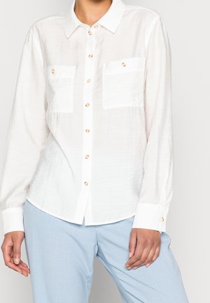 Hvid skjorte med lange ærmer og knapper med to brystlommer, kombineret med lyseblå bukser på en stående person.