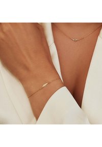 Gouden armband en ketting met een bladontwerp, gedragen op een huidkleurige arm en borst; beide hebben delicate kettingen en een gladdere textuur.