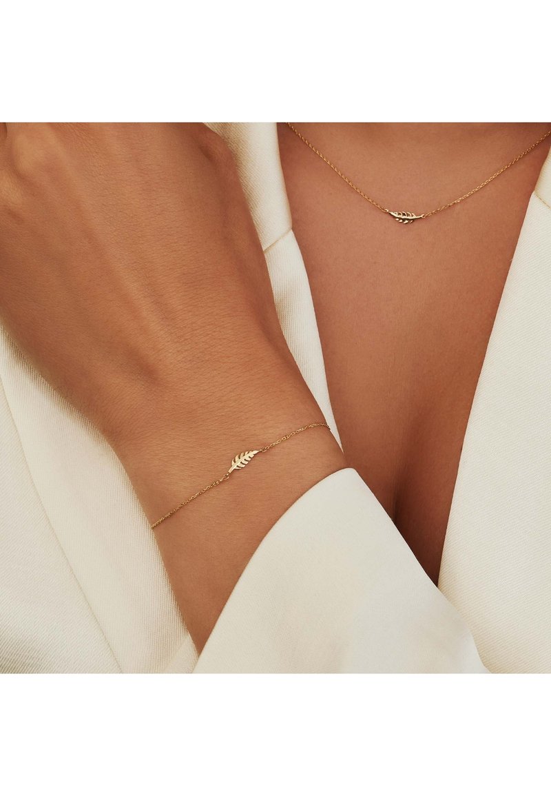Gouden armband en ketting met een bladontwerp, gedragen op een huidkleurige arm en borst; beide hebben delicate kettingen en een gladdere textuur.