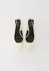 Ballerines slingback pointues blanches avec semelles intérieures noires rembourrées et nœuds géométriques décoratifs sur les orteils, présentées sur un fond blanc.
