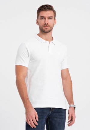 Vit bomullspolo med krage, kort ärm och en liten logotyp på bröstet. Bärs med mörkblå jeans.