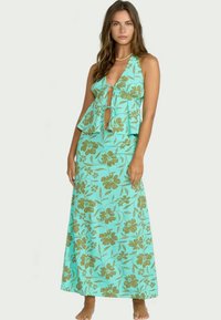 Femme portant une robe maxi turquoise à bretelles avec un imprimé floral marron, dotée de découpes à l'avant et d'un détail en peplum fluide à la taille.