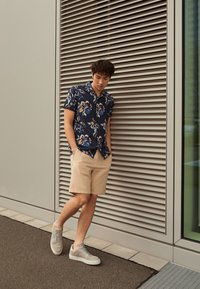 Camisa de manga corta azul marino con estampado floral, pantalones cortos beige y zapatillas gris claro, de pie contra un fondo arquitectónico moderno.
