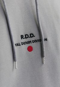 Šedá mikina s kapucí s černou výšivkou "R.D.D. Royal Denim Division". Obsahuje červený kruh a šňůrky se stříbrnými konci. Měkká textura látky.