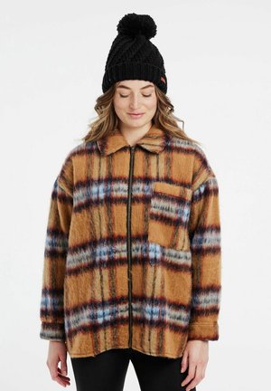Bruin geruite fleece jas met blauwe en rode strepen, voorzien van een rits aan de voorkant, een kraag en een borstzak. Draagt een zwarte gebreide beanie.