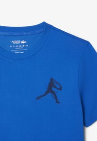 Sininen puuvillainen t-paita, jossa on pyöreä kaula-aukko. T-paidassa on rinnan kohdalla pelaajan siluetti ja kohopainettu motivoiva teksti kauluksessa.