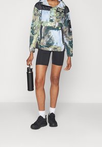 Pulloverjacka med blå och grön skogsprint, svarta accenter, elastiska ärmslut; kombinerad med svarta shorts och svarta sneakers, håller en svart vattenflaska.