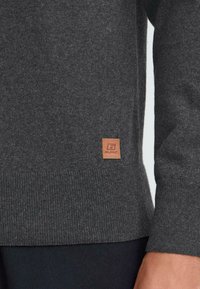 Maglione in maglia grigio scuro con orlo e polsini a coste, caratterizzato da un piccolo patch in pelle marrone con il logo "Blend" vicino alla vita, indossato sul lato destro.
