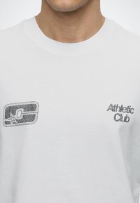 Κοντινό πλάνο λευκού T-shirt με ξεθωριασμένο μαύρο κείμενο "Athletic Club" στο δεξί στήθος και φθαρμένο ορθογώνιο λογότυπο με αστέρι στο αριστερό στήθος.