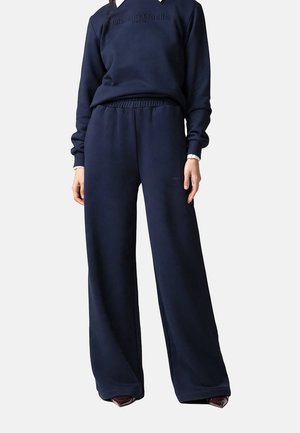 Persona que lleva pantalones anchos azul marino y sudadera a juego con un sutil logo en relieve, combinados con zapatos oscuros y puntiagudos.